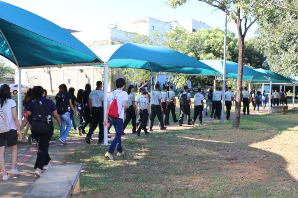 Grupo de estudantes caminha por uma área arborizada do Câmpus de Palmas, seguindo por uma passarela coberta por estruturas azuis. Os participantes estão em fila durante visita guiada, cercados por gramado, árvores e prédios universitários ao fundo.